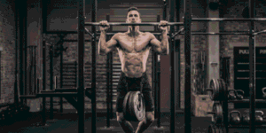 A muscular man in a gym doing a weighted pull-up on a metal rig