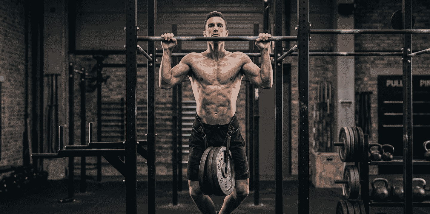 A muscular man in a gym doing a weighted pull-up on a metal rig