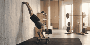 Athlete performing a wall-assisted handstand push-up on parallettes in a gym