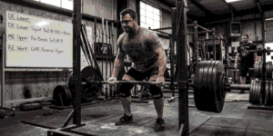 A male athlete deadlifting a heavily loaded barbell in a gym, wearing a belt and knee sleeves, with a whiteboard of workout instructions in the background
